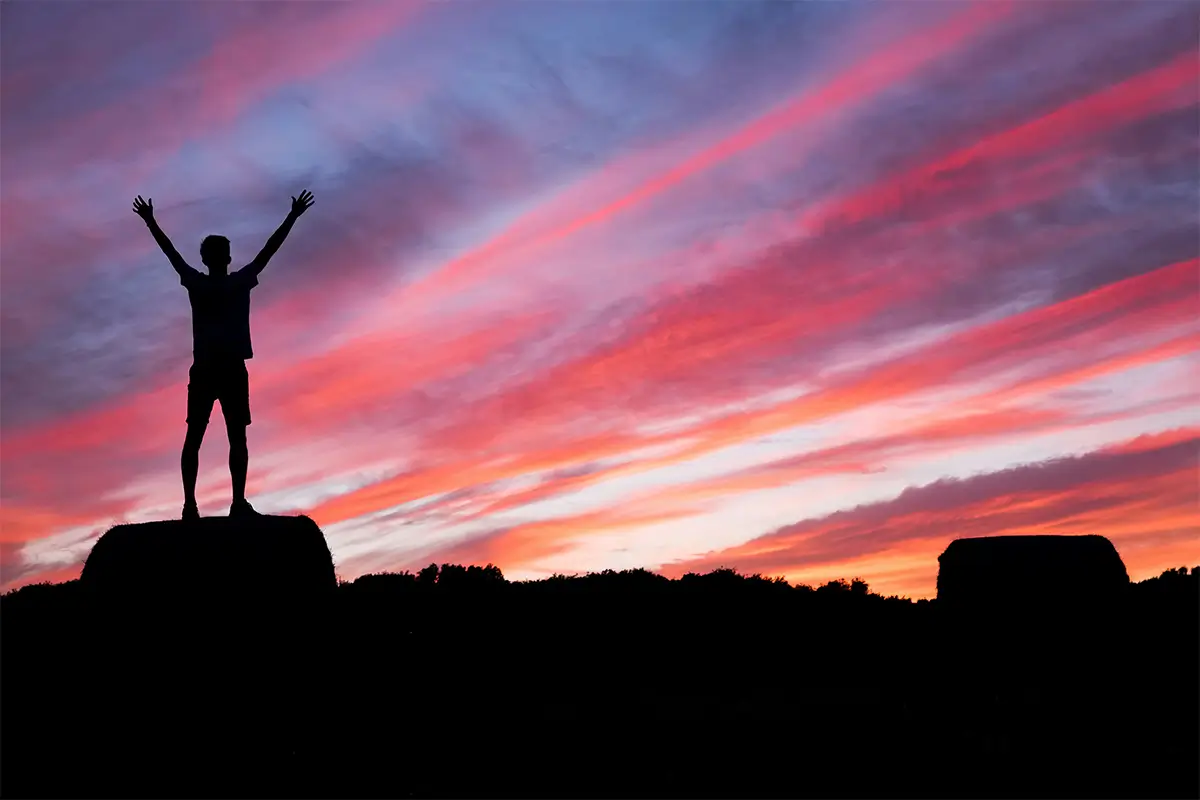 A silhouette of a man raising his hands while standing on a rock at sunset
