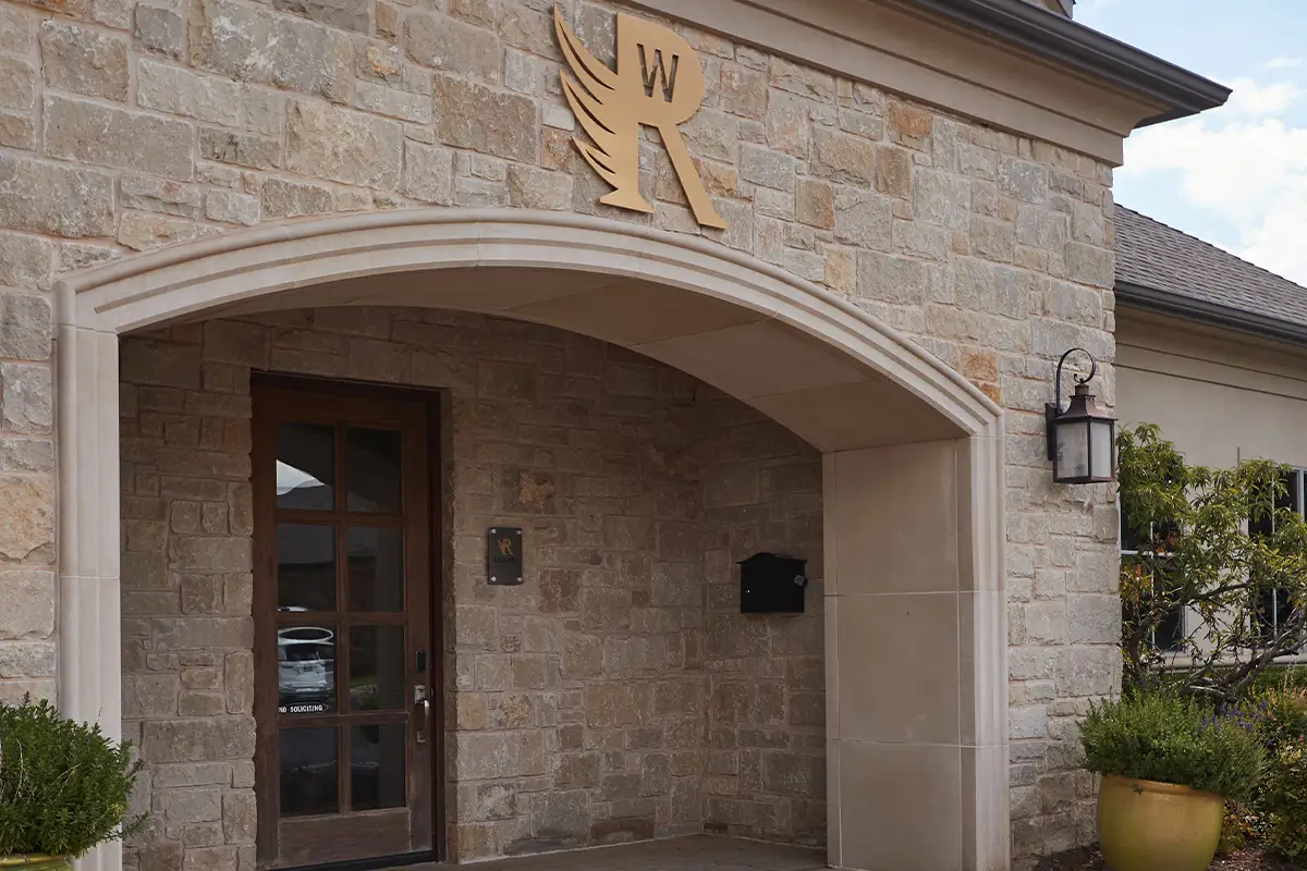 Stone exterior entrance with arched entryway, wooden glass-paneled door, and decorative wall lantern at a residential or clubhouse-style building.