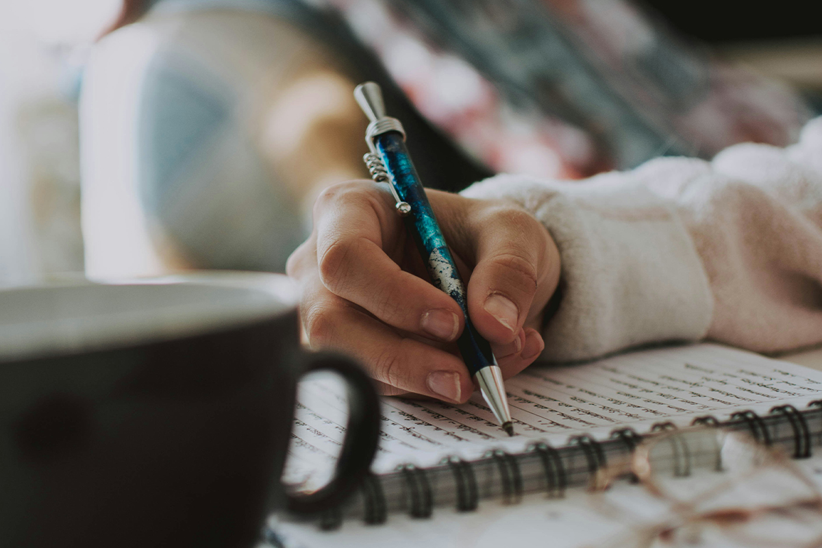 An extreme close-up of a person's hand holding a blue and silver patterned pen, poised to write in a spiral-bound notebook.