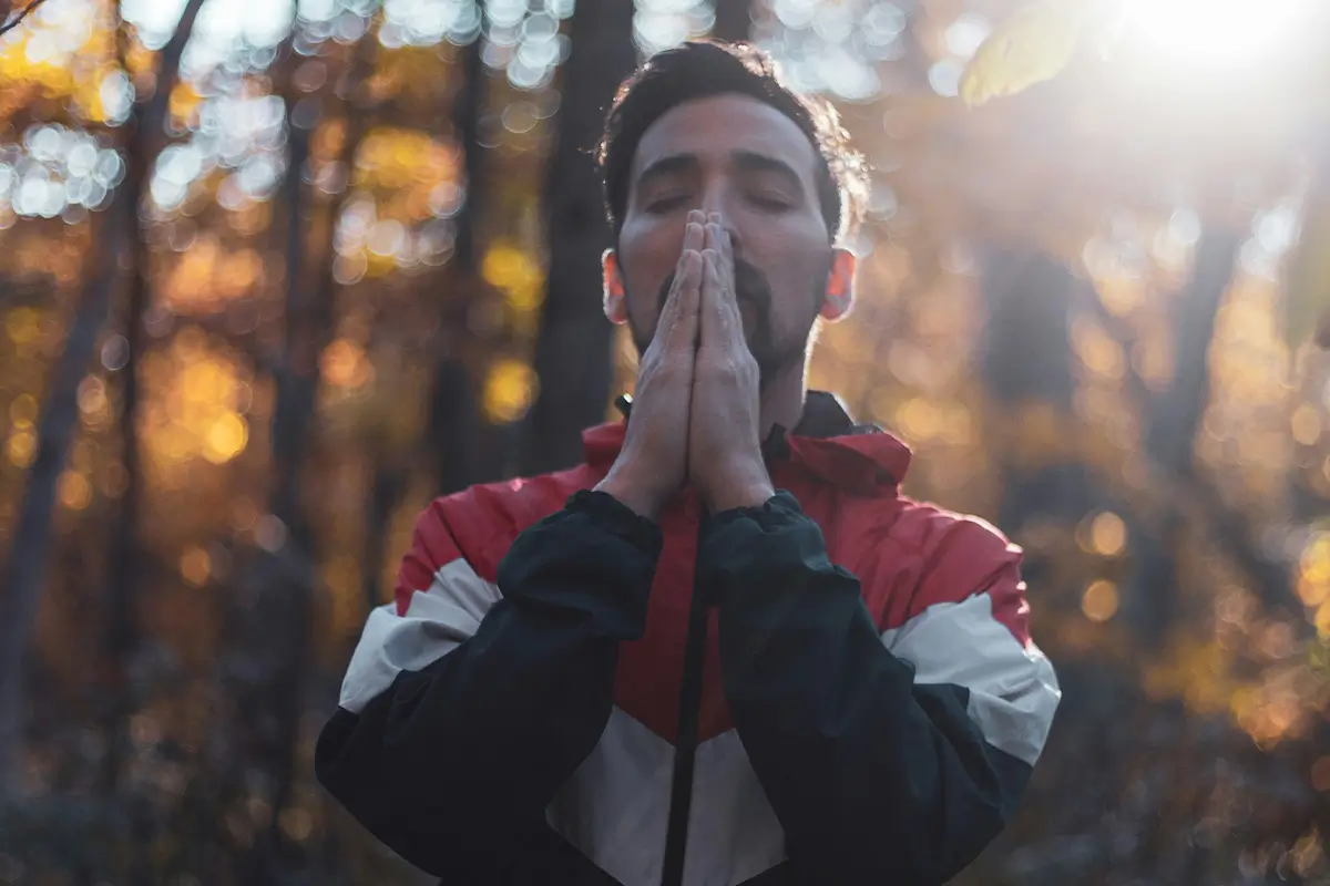 A man with closed eyes and hands pressed together in prayer stands in a sunlit forest with autumn foliage.