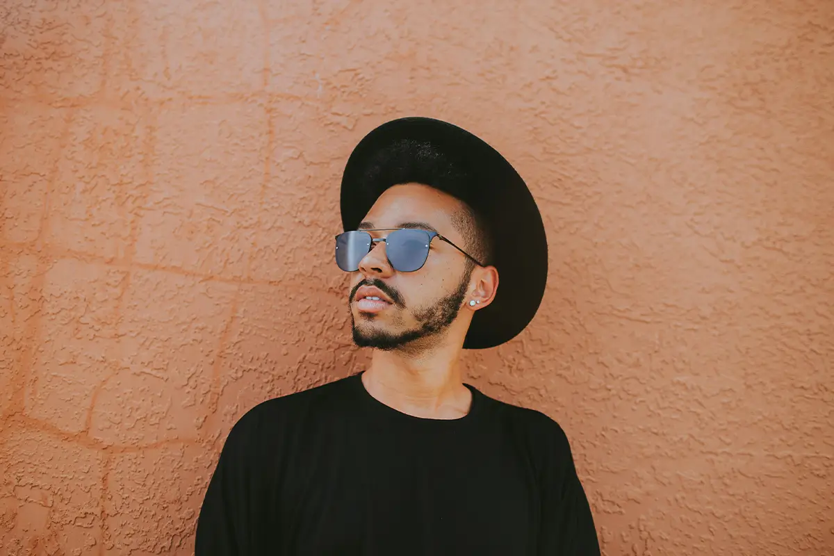 The Mind-Body Connection: Chronic Pain and Depression Man wearing a black hat and sunglasses standing against a textured terracotta wall.